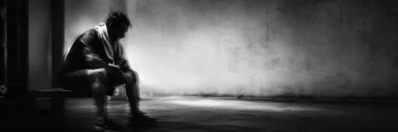 A man sits on a bench in a dimly lit room, wearing a towel around his neck, appearing contemplative or exhausted.