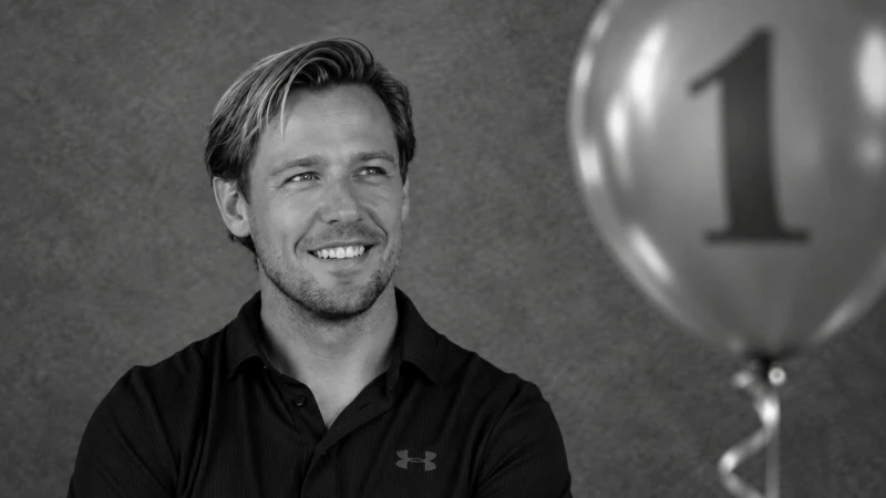 Een lachende man in een zwart shirt kijkt naar links. Rechts is een ballon met het cijfer 1 zichtbaar. Grijze achtergrond.