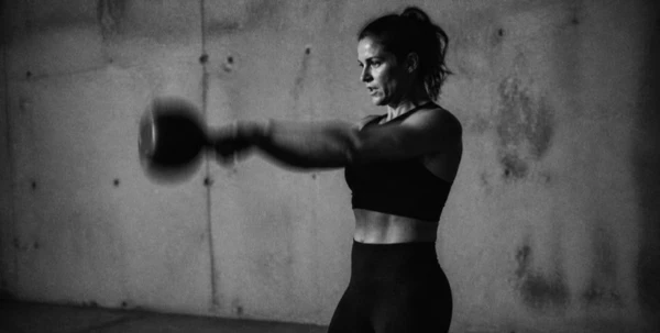 Woman in workout attire swinging a kettlebell in a dimly lit gym.