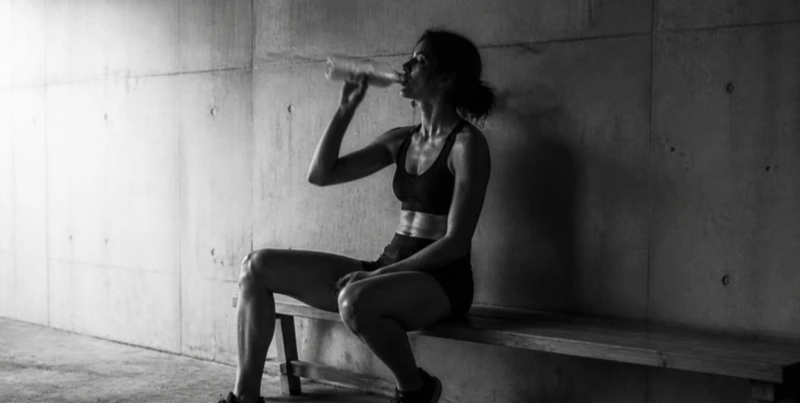 A woman in workout attire sits on a bench against a concrete wall, drinking from a bottle in a dimly lit space.