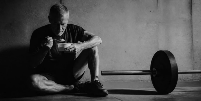 Man sitting on the floor eating from a bowl, next to a barbell, in a dimly lit room.