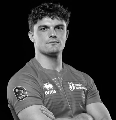 Een man in een Rugby Nederland-shirt met gekruiste armen, kijkt serieus naar de camera tegen een zwarte achtergrond.