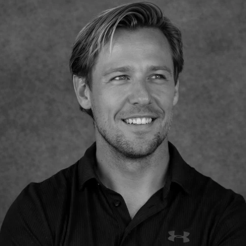 Een lachende man - Ian van der Werf - met kort haar in een zwart poloshirt, kijkt naar rechts. Grijze achtergrond. 