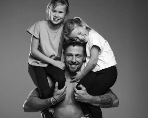 Een man draagt twee lachende kinderen op zijn schouders, in een zwart-wit foto. Ze lijken gelukkig en ontspannen.