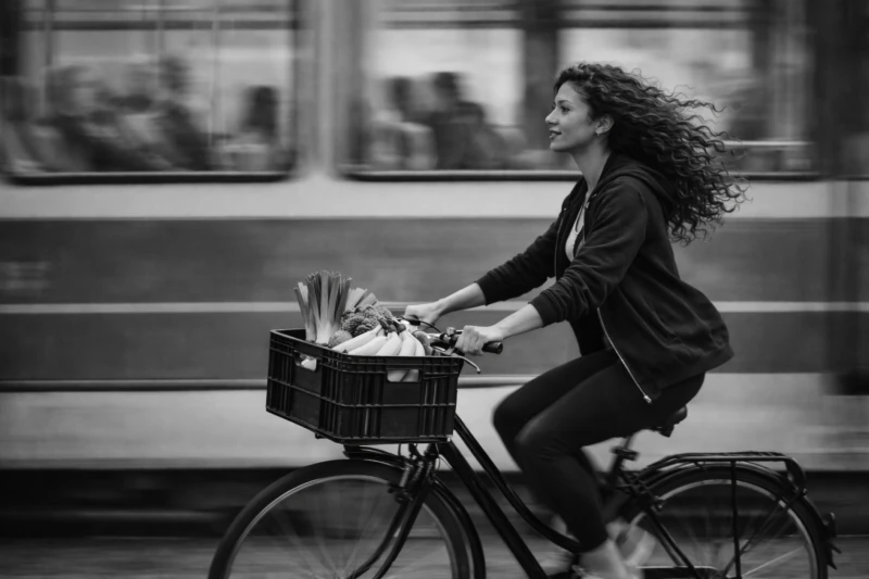 Een vrouw fietst met een mand vol groenten voorop, langs een voorbijrijdende tram.