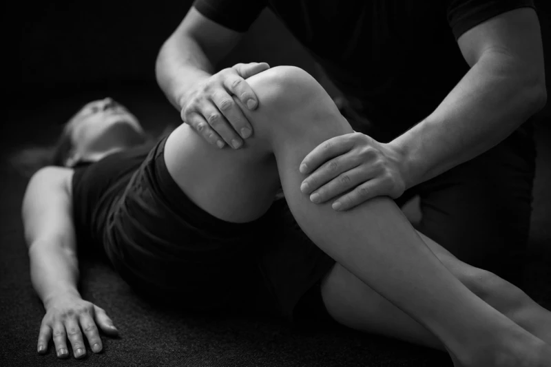 Een persoon ligt op de grond, een ander houdt hun gebogen knie vast, mogelijk tijdens fysiotherapie. Zwart-wit foto.